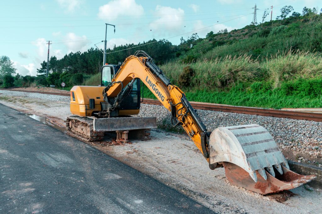 Yellow excavator parked on a construction site beside railroad tracks and greenery.