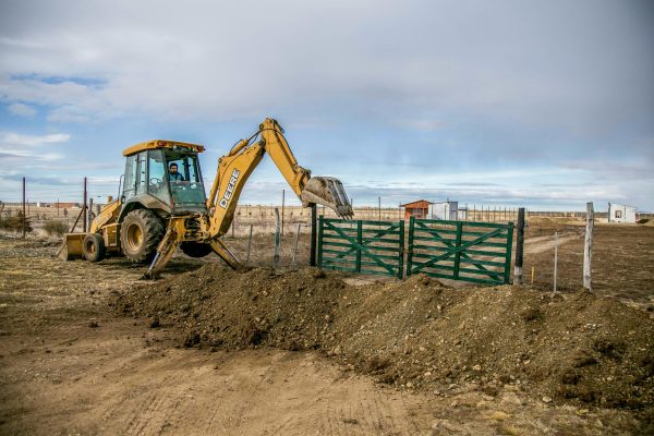A yellow backhoe loader actively digs soil at a farm entrance with a green gate, under a cloudy sky.